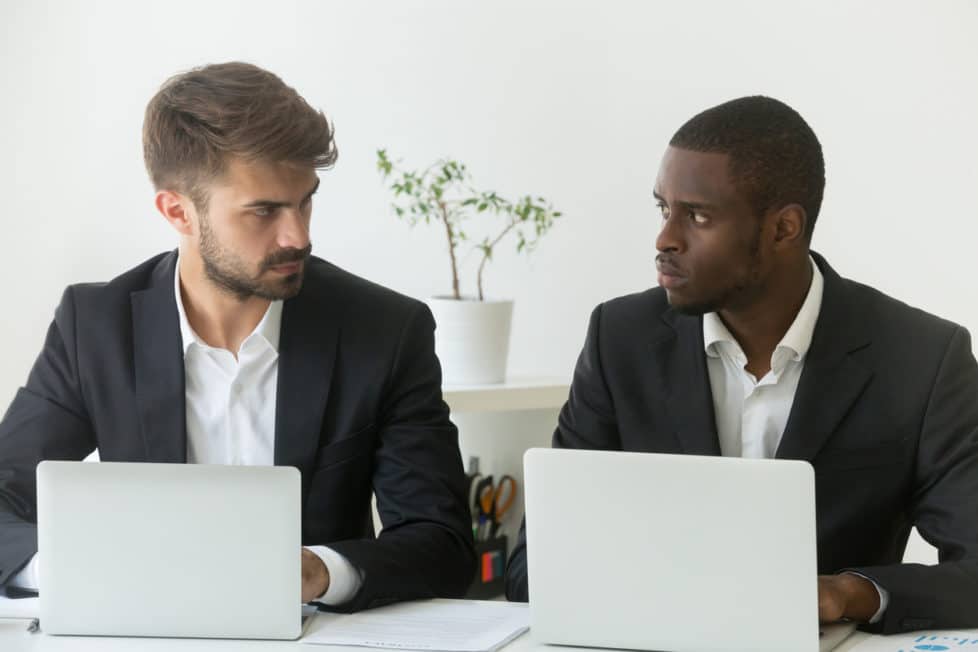 Zwei Kollegen sitzen nebeneinander am Laptop und beäugen sich kritisch - Konkurrenzkampf Jobfrust