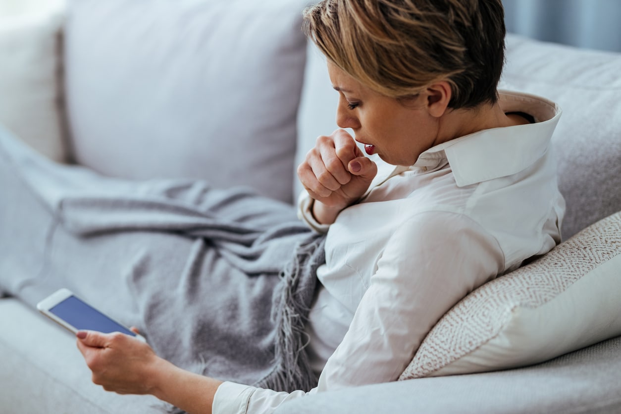 Kranke Frau auf Couch hustet und hält Handy für Krankmeldung in der Hand